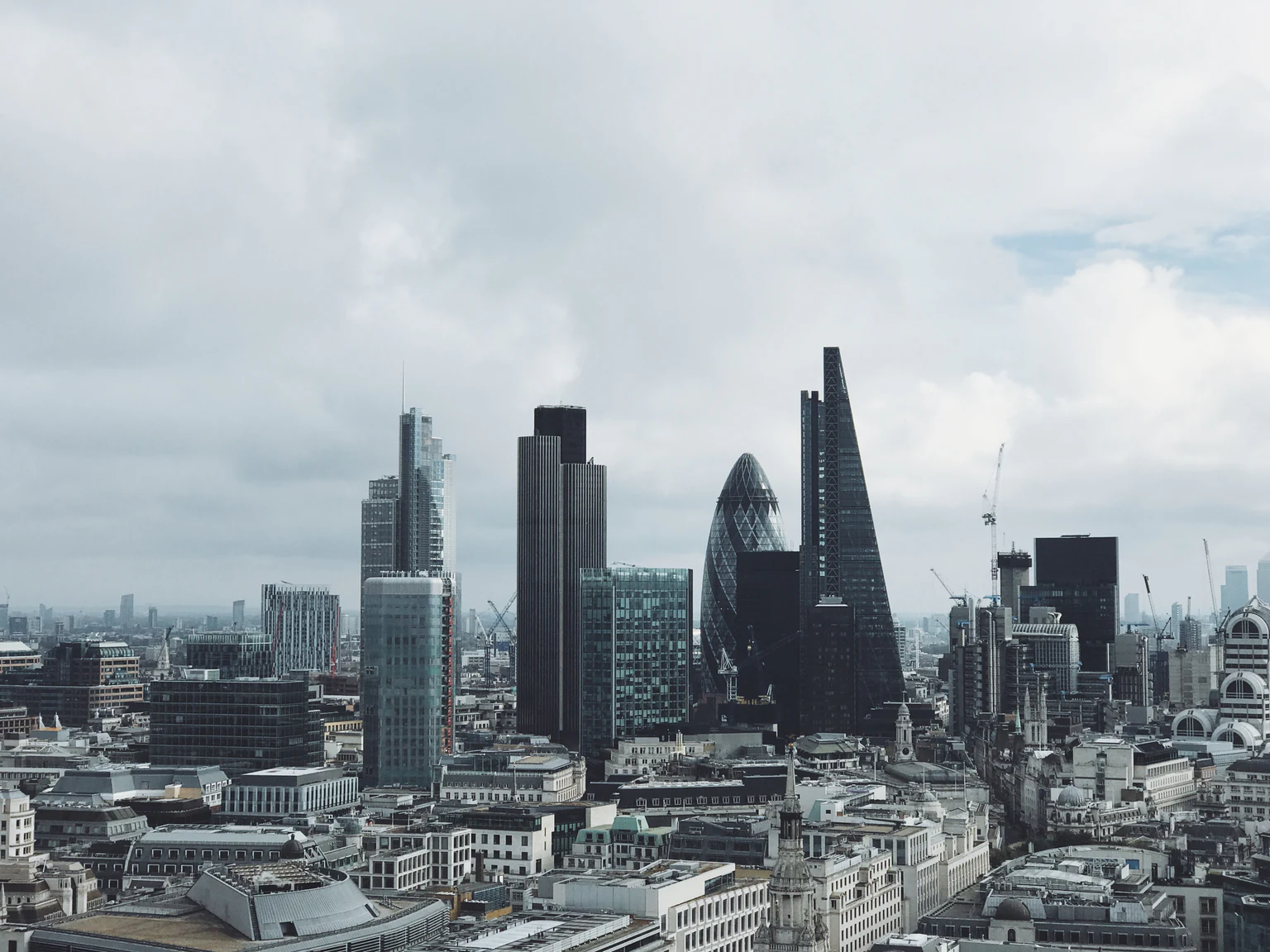 City of London rooftops with the Gherkin and Leadenhall Building beneath a cloudy sky.
